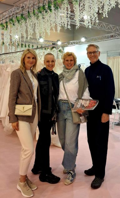 2026 European Bridal Week - Meeting people - Team Braut und Braeutigam - Volker Rohde Vier Personen stehen lächelnd in einem Festsaal mit Hochzeitskleidern und Dekoration.